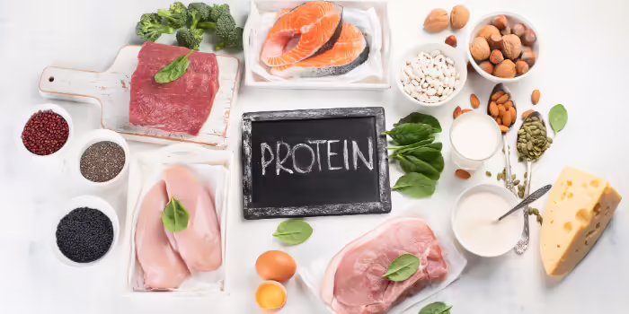A variety of high‑protein foods arranged on a white surface and a small chalkboard in the center displays the word ‘PROTEIN.