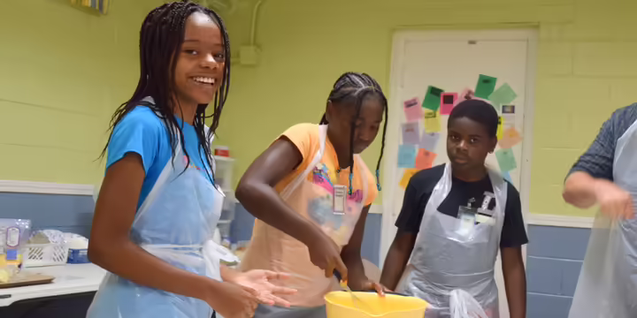 Children with aprons; one is stirring food in a bowl.