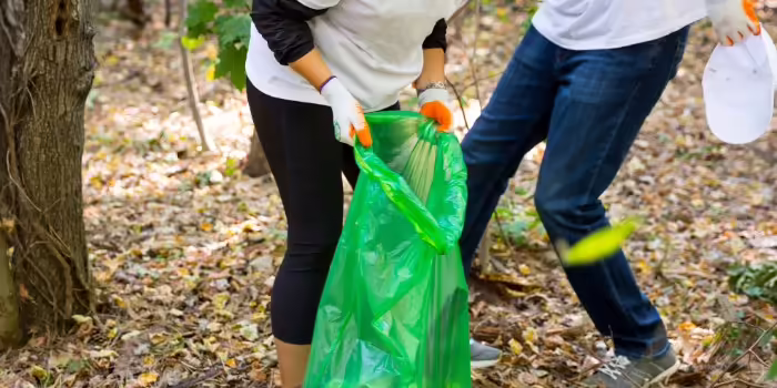 Two people pick up trash in the forest with gloves and a large green bag.