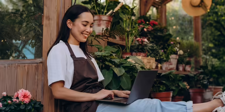 Woman typing on laptop in a garden setting with a garden apron on. 