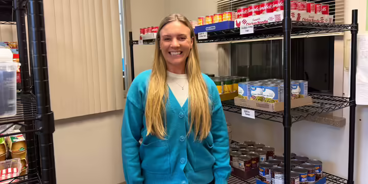 Morgan is pictured in a blue jacket, standing in front of a food pantry display
