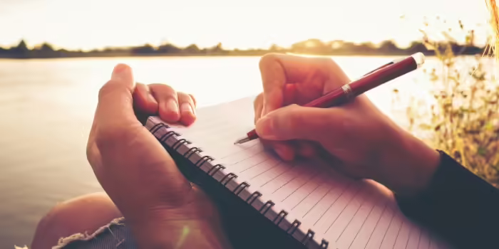 A person writes in a notebook by a lake. 