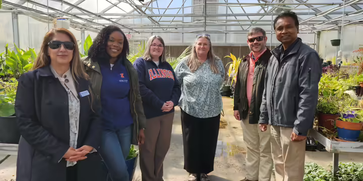 Six people, some wearing branded University of Illinois apparel, stand inside a greenhouse