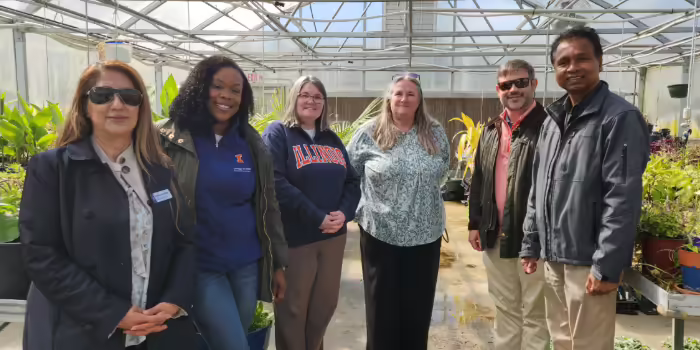 Six people, some wearing branded University of Illinois apparel, stand inside a greenhouse