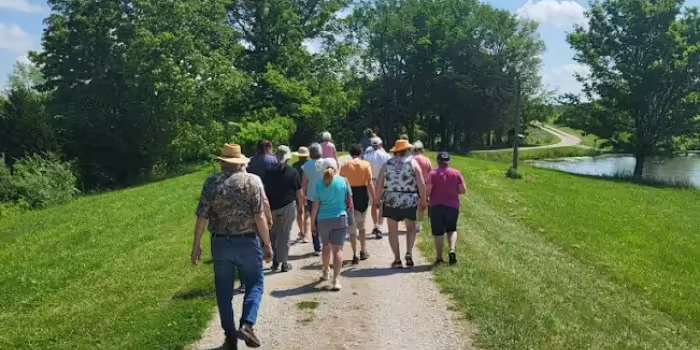 Group of adults walking together along a gravel path in a sunny park, with green grass, trees, and a pond beside a winding road in the background.