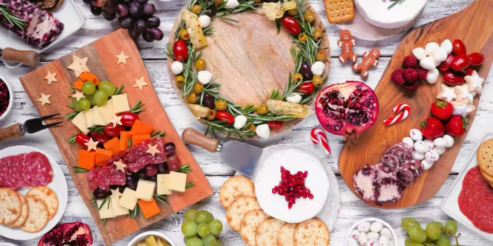 A festive holiday charcuterie spread displayed on wooden boards arranged in shapes such as a Christmas tree and a wreath, set on a rustic white wooden table.