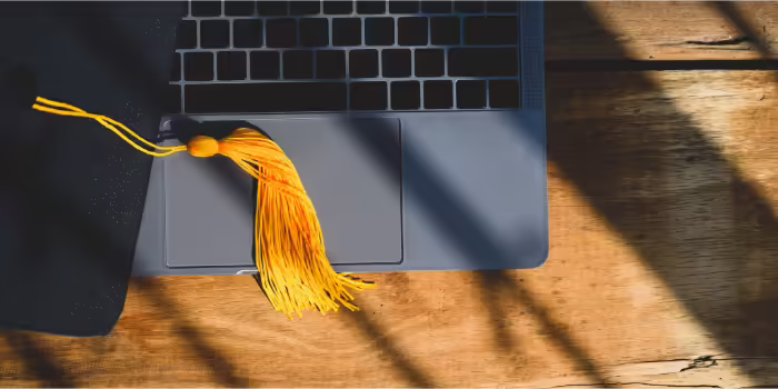 A black graduation cap with a yellow tassel resting on the trackpad of a laptop. The laptop is on a wooden surface, with sunlight casting shadows across the scene.
