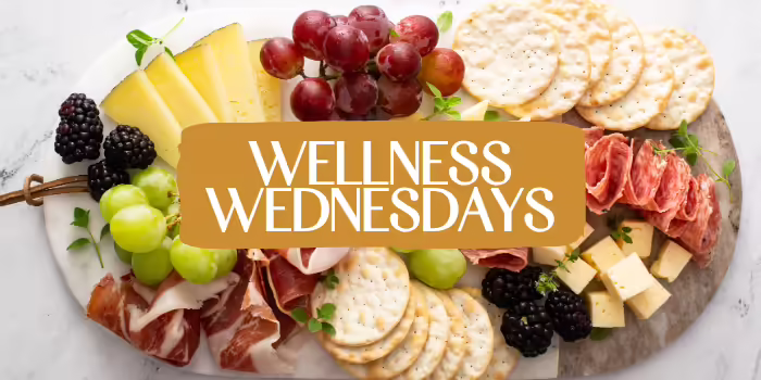 An assortment of cheeses, crackers, grapes, blackberries, and sliced meats arranged on a charcuterie board with the text “Wellness Wednesdays” in the center.