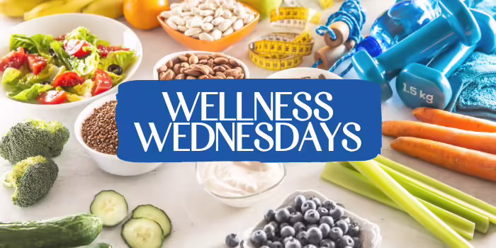 A variety of healthy foods and wellness items displayed on a table, with a blue banner in the center that reads “Wellness Wednesdays.”