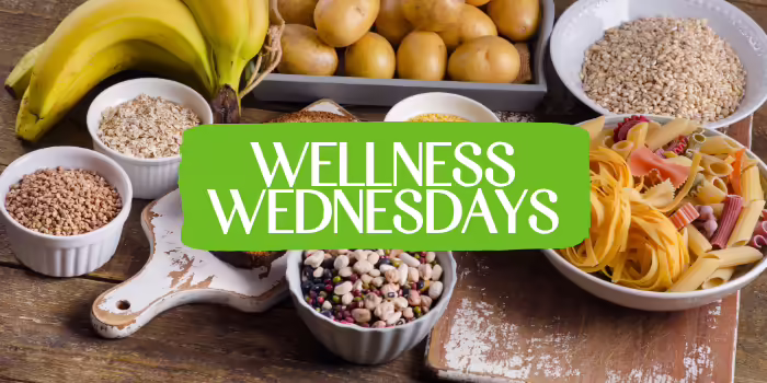 Wellness Wednesdays text centered on an green banner over a background of fresh vegetables and healthy ingredients.