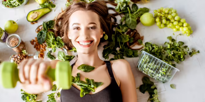 A smiling woman in workout clothing holds a green dumbbell while surrounded by fresh fruits, vegetables, leafy greens, and other healthy foods.