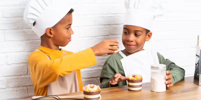 Two children wearing white chef hats and aprons sit at a table with decorated cupcakes.