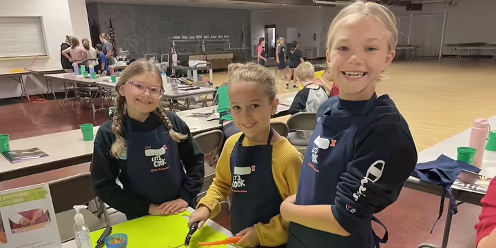 Three girls wearing cooking aprons in a kitchen setting and peeling carrots. There are lime green cutting boards and knives in front of them.