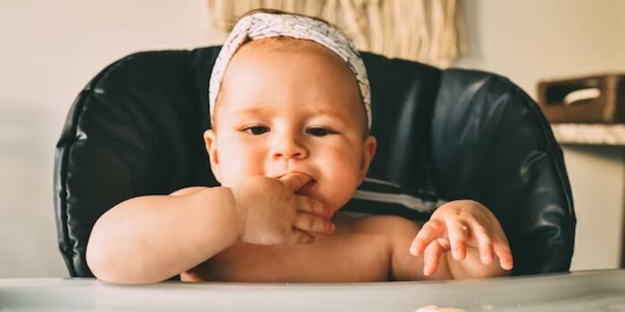 Light-skinned baby with a bow sitting in a highchair reaching for a cracker.