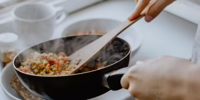 Someone holding a black pan, scrapping food into a bowl with a wooden spoon.