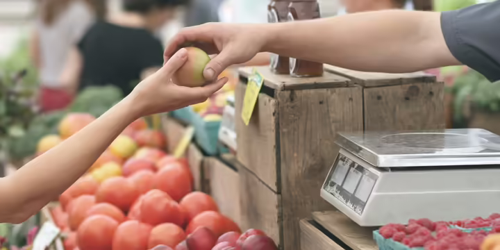 A display fruits and vegetables. There is a person giving another person an apple. 