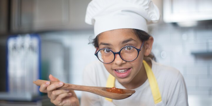 A young girl holding a spatula. She is wearing a hat and glasses.