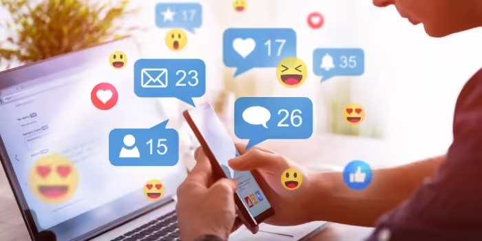 a person holding their phone with their laptop open also and different emojis and notifications floating around