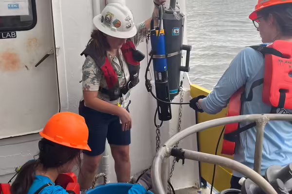 Three women wearing hard hats and flotation devices in a boat use equipment to measure water quality