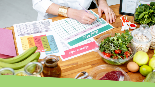 A person sitting at a wooden table filling out a meal planning chart with various fruits and vegetables sitting around the chart on the table.