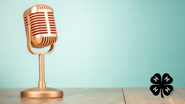 A gold microphone sitting on a wooden desk with a blue background. A black 4-H clover in the bottom right corner.