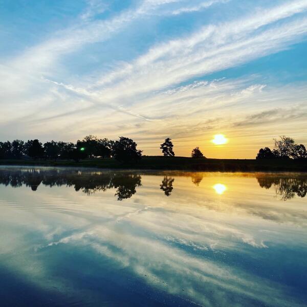 a still pond at sunrise with the reflection of the sky, clouds and trees on the water