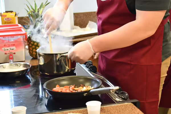 A pair of hands stirring a pot as a pan of sausages fries in the foreground.