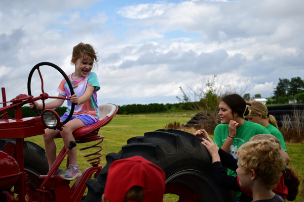 A girl sitting in the driver's seat of a tractor as others gather around the tractor.