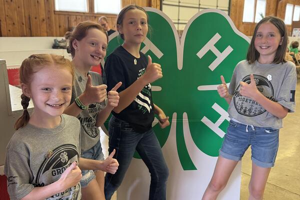 4 kids with thumbs up in front of a large 4-H clover