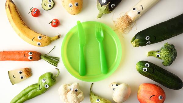 Child's empty plate surrounded by fruits and vegetables wearing googly eyes