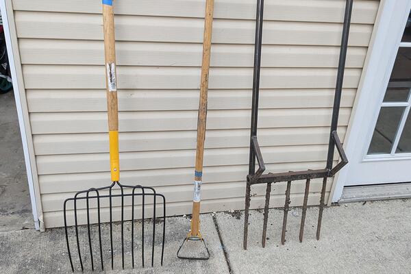 A row of garden tools including a silage fork, stirrup hoe, and broadfork all leaning against a house. 