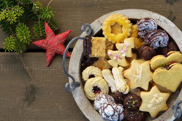 A plate filled with a variety of holiday cookies of shapes, colors, and flavors sitting on a wooden table next to evergreen holiday décor.