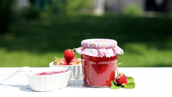 A jar of strawberry jam sits on a table with a bowl of strawberries.