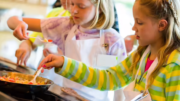 two young girls cooking food in a pan and tasting the food with a spoon