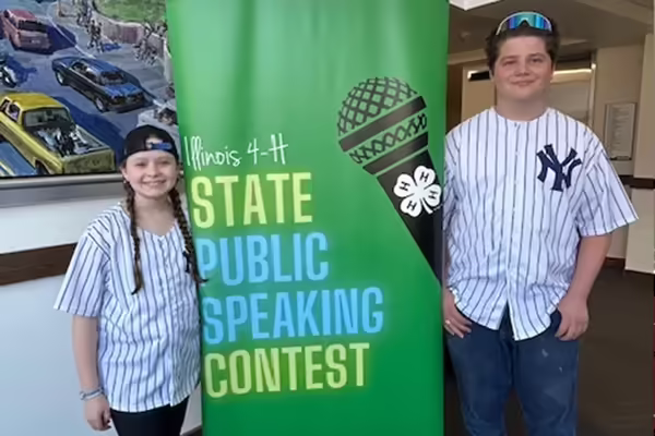 2 youth wearing baseball jerseys standing in front of a banner that says "Illinois 4-H State Public Speaking Contest"