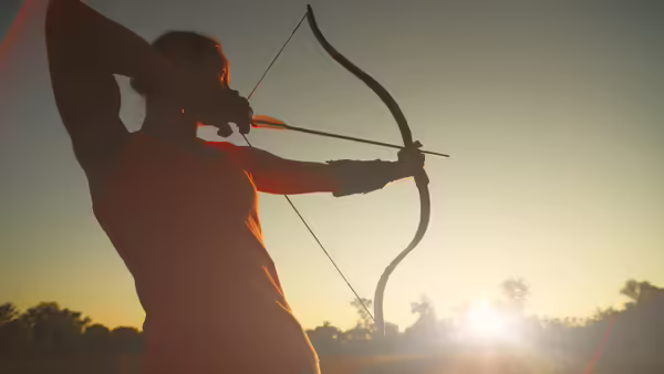 female archer shooting with a bow in a field at sunset