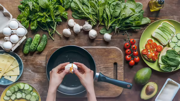 A person cracking an egg into a frying pan, surrounded by various ingredients to make a sandwich.