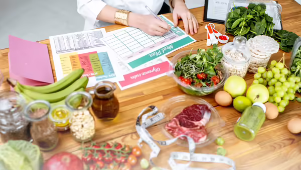 A person sitting at a wooden table filling out a meal planning chart with various fruits and vegetables sitting around the chart on the table.