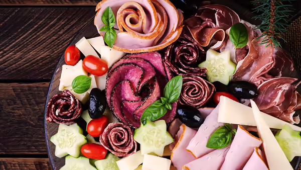 A charcuterie board with an assortment of meats, cheeses, and garnishes. The board is arranged on a dark wooden surface.