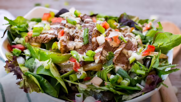 A steak salad in a white bowl, garnished with chopped green onions and diced tomatoes, and drizzled with a creamy white dressing. The bowl sits on a white background with a wooden surface.