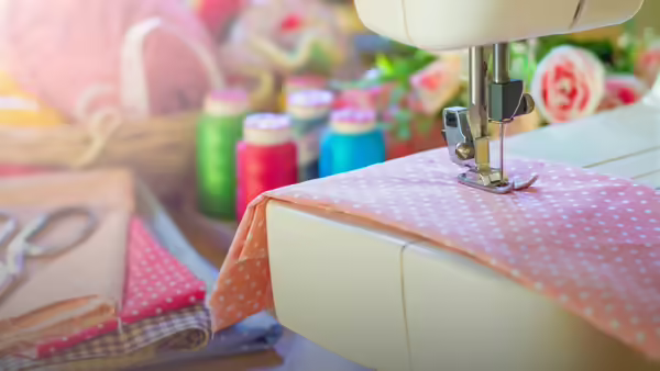 A sewing machine stitching a pink, polka-dot fabric, with colorful thread spools and scissors in the background.