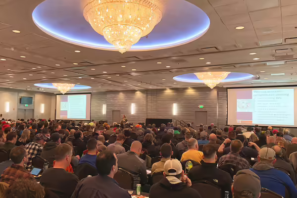A large room completely filled with people sitting in chairs at tables listening to a training presentation.