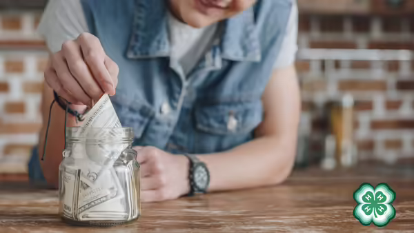 A person is placing folded dollar bills into a clear glass jar. A green 4-H clover in bottom right corner.