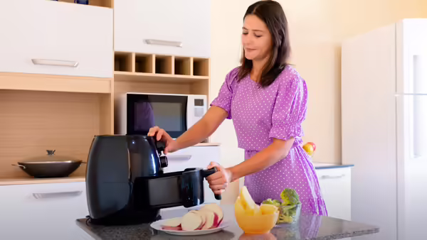 A woman in a purple dress places food into a black air fryer on a kitchen counter.