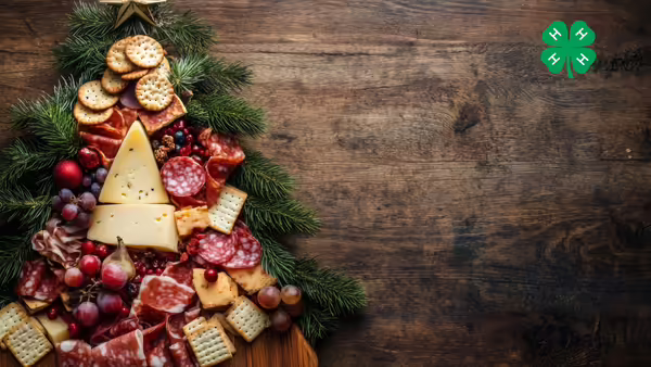 A charcuterie board in the shape of a tree with cheese, meats, crackers a wooden background. A green 4-H clover is in the top right corner.