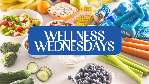 A variety of healthy foods and wellness items displayed on a table, with a blue banner in the center that reads “Wellness Wednesdays.”