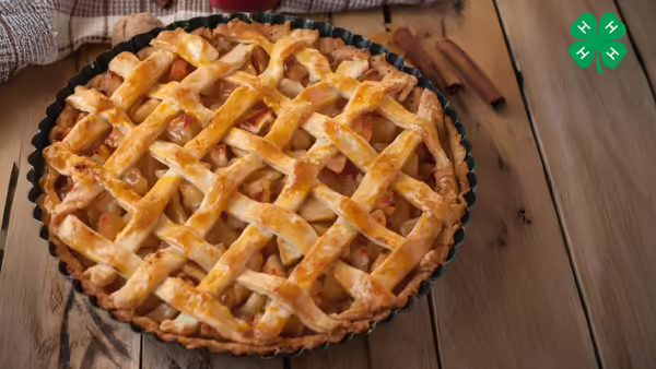 A baked apple pie with a golden-brown lattice crust in a metal pan on a wooden table. The green 4-H logo is in the right upper corner.