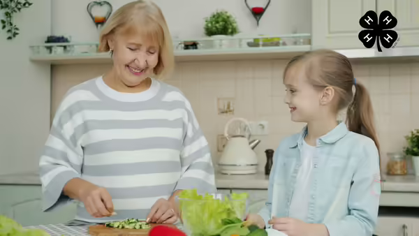 A grandmother and granddaughter in a kitchen. The grandmother chops vegetables on a cutting board. The black 4-H logo is in the upper right corner.