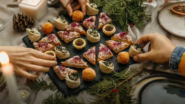  Hands reach for an assortment of appetizers arranged on a dark platter set on a table decorated with evergreen and candles.