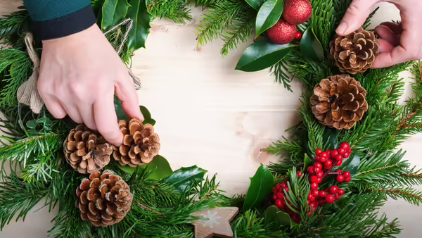 Hands arranging pinecones and holly berries onto a holiday wreath made of evergreen branches